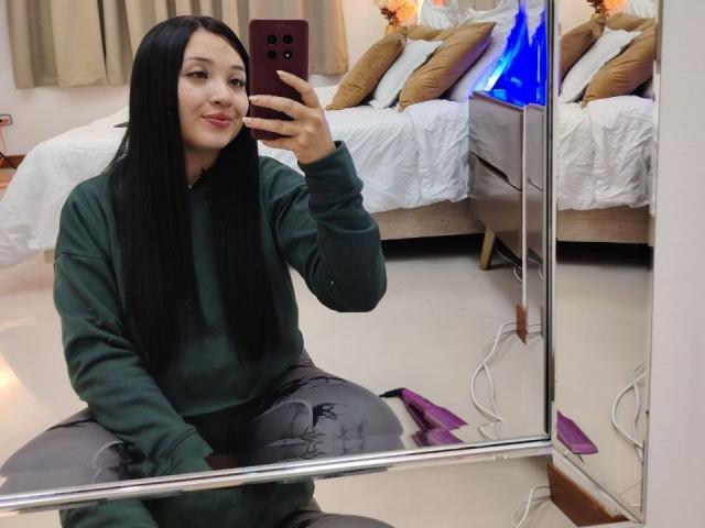 LunaBellaHotty, with long black hair, green hoodie, and gray pants, kneeling, taking a mirror selfie with a pink phone, smiling, in a bedroom with a blue lamp.