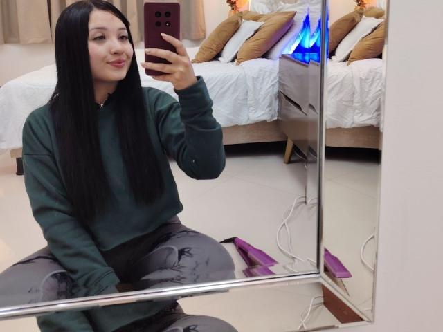 LunaBellaHotty with long black hair, green sweater, and gray pants, kneeling, taking a mirror selfie with a purple phone, blue laptop light on bed, purple vibrator on floor.