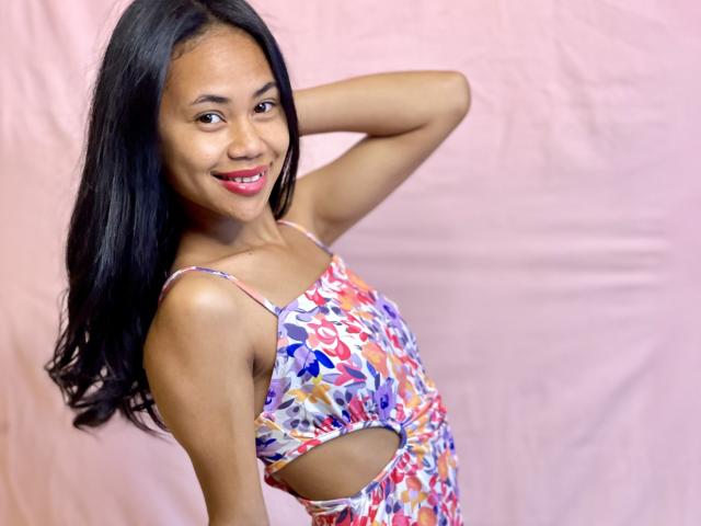 AubreyLoo with long black hair, smiling, wearing a colorful, cut-out dress, has small tits, and a cute, cute ass, against a pink backdrop, raising one arm.