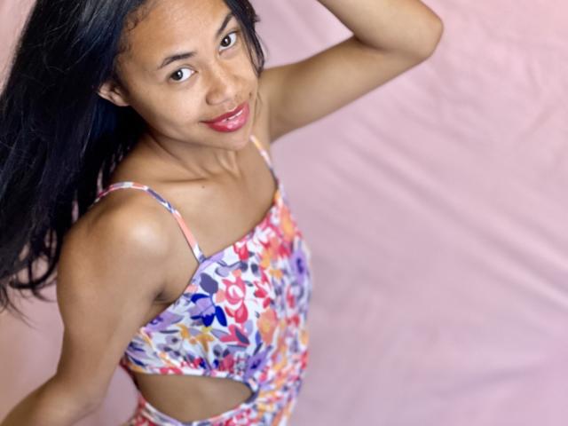 AubreyLoo with dark skin and long black hair, wearing a colorful floral dress, smiling at the camera, showing small tits and a cut-out midsection, arm raised.
