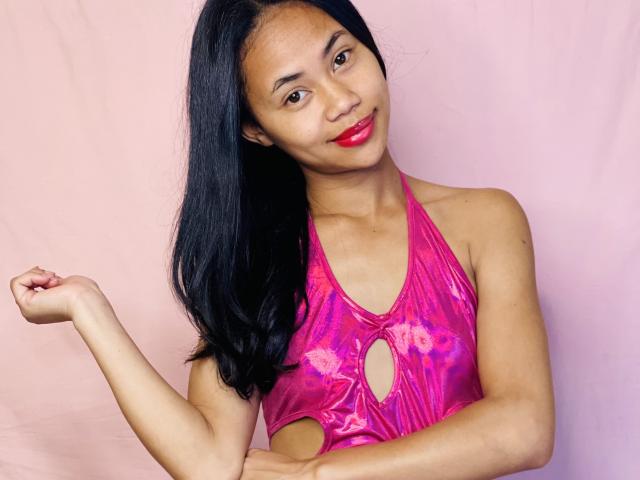 AubreyLoo with long black hair, red lipstick, and pink metallic halter top with cutout, raising her right arm, smiling, showing her small, cute tits against a pink background.