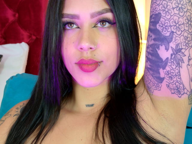 AgathaVelez69, com cabelos longos e pretos, sombra roxa, batom rosa, piercing no nariz, piercing no lábio, tatuagens no braço direito e uma pequena tatuagem no pescoço, sob luzes roxas e amarelas..