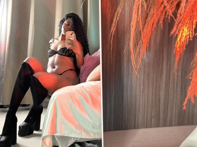 GianellaArittza with dark skin, curly hair, black lace bra, black thong, black thigh-high boots, sitting on bed, holding phone, showing her big ass and tits. Right side: orange fern wall.