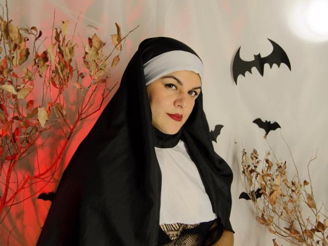 AlissaBels with pale skin, red lipstick, black and white nun outfit, black veil, and fishnet top, smiling at camera, surrounded by bat decorations and dry branches.