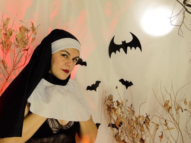 AlissaBels in a nun outfit with a black veil and white wimple, red lipstick, and black dress, surrounded by bat cutouts and dry branches, under a bright light.