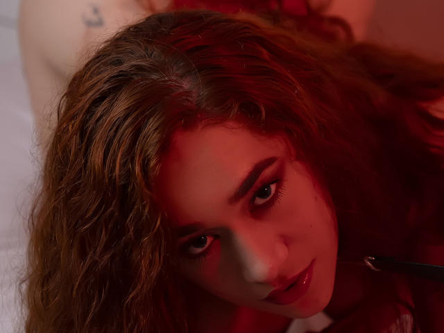 ValeryHilton has curly brown hair, red-lit face, and dark eyes, looking seductively at the camera with slightly parted, shiny red lips. Her bare shoulder is visible.