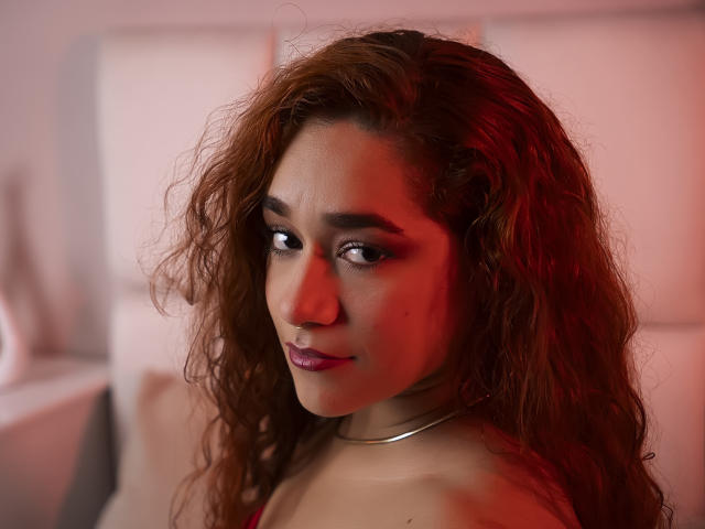 ValeryHilton has curly brown hair, dark eyes, and purple lipstick. Red lighting highlights her face. She's wearing a metallic choker and a red top, looking slightly annoyed.