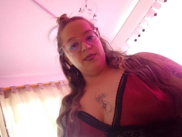 CleareRems with long brown hair, glasses, red sheer top showing black lace trim, and a chest tattoo, smiling slightly. Pinkish light from above. Boobs visible.