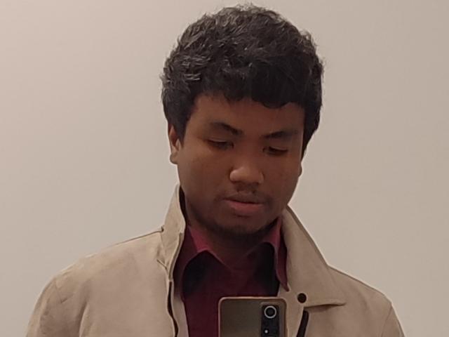 HottManX with short curly black hair, brown skin, looking down, wearing a beige jacket over a red shirt, holding a phone in front of him.