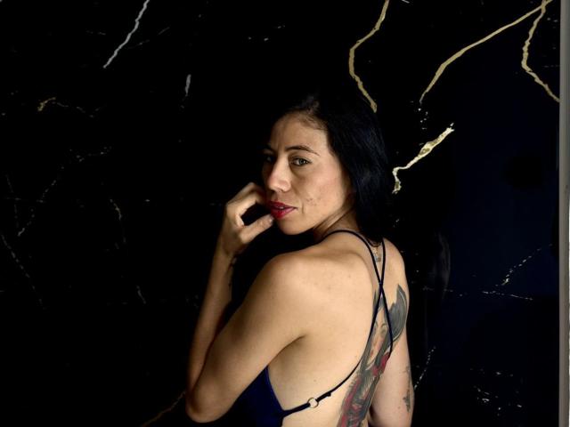 KattySloven, avec des cheveux noirs, regardant en arrière, portant une robe bleue, montrant un tatouage sur son dos, mordant son doigt, fond noir avec des fissures dorées..