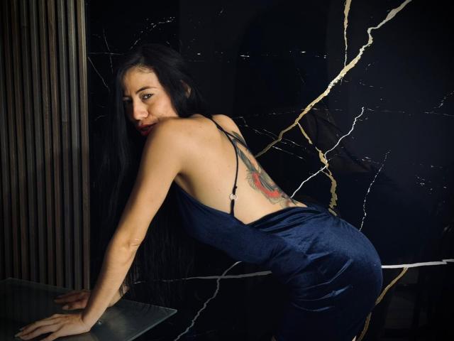 KattySloven, con lunghi capelli neri, indossa un abito di seta blu, è piegata in avanti, mostrando un tatuaggio sulla schiena, con le mani su una superficie di vetro, e guarda oltre la spalla..
