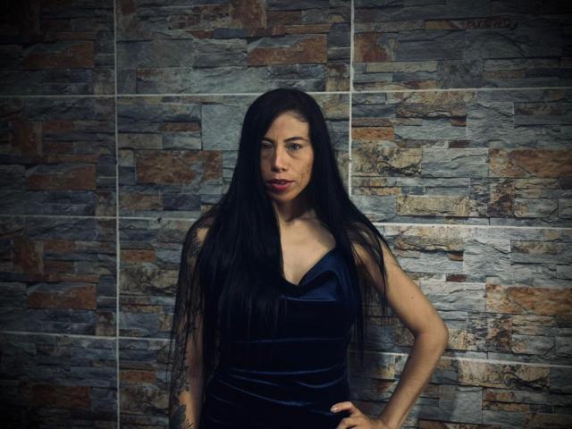 KattySloven with long black hair, wearing a dark blue velvet dress, standing against a stone wall, hands on hips, looking slightly annoyed, with a subtle smirk on her face.