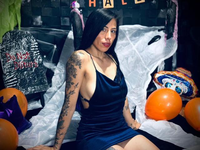KattySloven sits in a dark, Halloween-themed setting. She has long black hair, a tattooed arm, and wears a low-cut, dark blue velvet dress. Orange balloons and a glittery sign are in the foreground. Her red lipstick contrasts with her dark dress. White plastic sheets are draped behind her, adding a spooky effect. She looks directly at the camera with a slight smile. The image uses sharp, vibrant colors and focuses on KattySloven's central position.