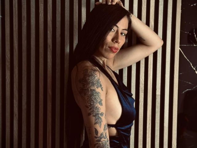 KattySloven, avec ses longs cheveux noirs, son rouge à lèvres rouge et sa robe en satin bleu, bras levé, montrant un tatouage détaillé sur sa manche droite, est sexy et confiante..
