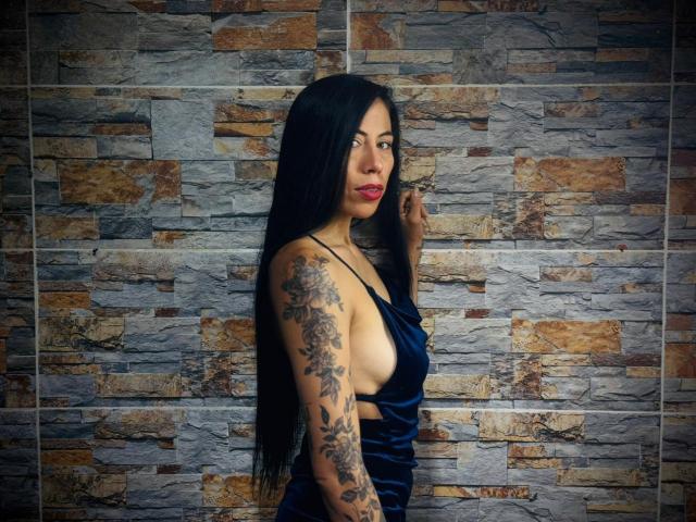 KattySloven with long black hair, blue velvet dress, sideboob, tattoos on right arm, red lipstick, standing against stone wall, holding phone to her ear.