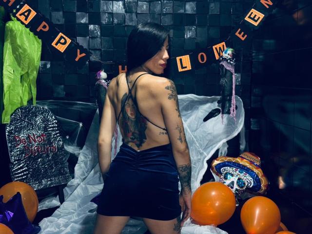 KattySloven mit Tattoos, mit dem Rücken zur Kamera, in einem schwarzen Samtkleid, das ihren Hintern zeigt, umgeben von Halloween-Dekorationen, orangefarbenen Ballons und einem Schädel. Dunkelhaarig, asiatisch, in einem schwach beleuchteten Raum..