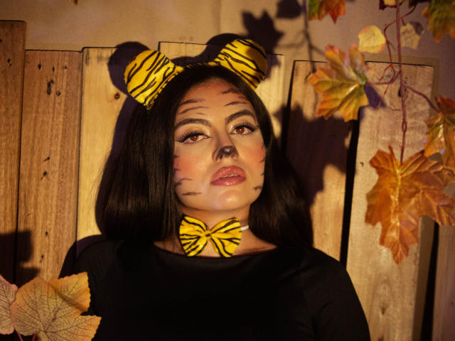 DorothyLane met een hoofdband, strik en gezichtsverf met strepen, zwart haar, gele ogen en glanzende lippen, gekleed in een zwarte top, omringd door herfstbladeren..