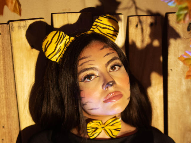 DorothyLane, con cabello negro, lleva orejas de tigre y un lazo, tiene bigotes falsos y labios brillantes, y mira con ojos marrones, con sombras en tablas de madera, bajo una luz cálida..