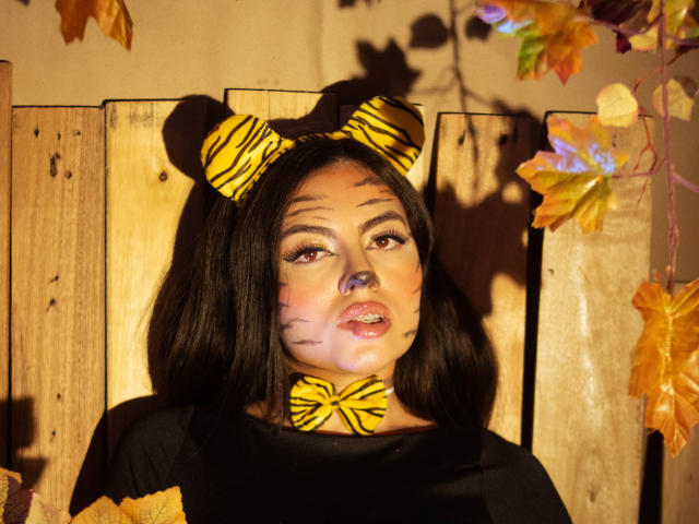 DorothyLane con orecchie da tigre e trucco sul viso, capelli neri, indossa una maglietta nera, foglie arancioni e gialle sullo sfondo, una recinzione di legno e una cravatta a farfalla gialla a forma di tigre..