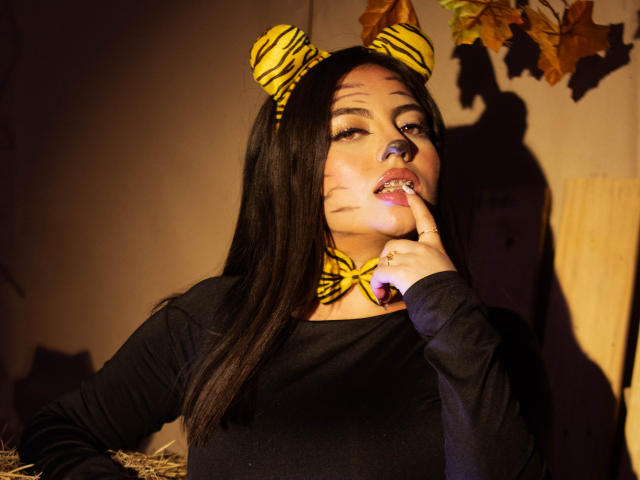 DorothyLane, com cabelo preto, usando orelhas de zebra e maquiagem no rosto, tem uma faixa de cabeça com listras amarelas, uma blusa preta e está chupando o dedo, com uma sombra na parede..