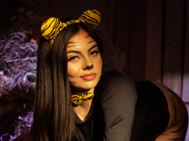 DorothyLane, con lunghi capelli neri, indossa una fascia per capelli gialla a righe di zebra e una collana coordinata, una maglia nera a maniche lunghe, è in posizione a quattro zampe, guarda la fotocamera, aspetto sexy, illuminazione calda, sfondo scuro..