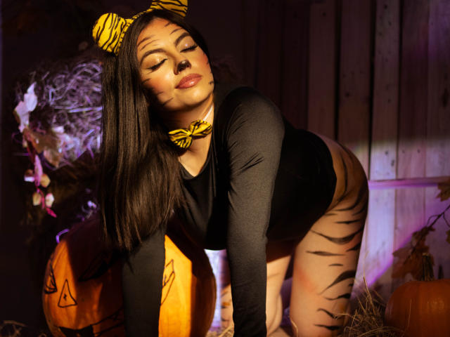 DorothyLane, con rayas de tigre en los muslos, una camiseta negra de manga larga, una corbata amarilla y orejas de cebra, inclinada hacia adelante, con los ojos cerrados, y con rayas de tigre pintadas en la cara..