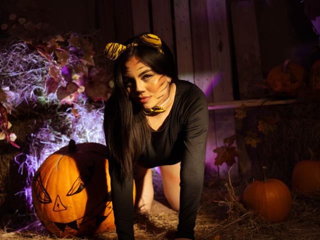 DorothyLane on all fours, wearing a black long-sleeve top, yellow cat ears, and a yellow necklace, with two carved pumpkins, one with a angry face, in the background.