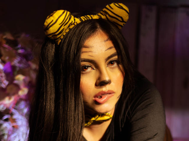 DorothyLane with long black hair, wearing tiger-striped headband and matching choker, has shiny pink lips and brown eyes, looking seductively at the camera. She's wearing a black top.