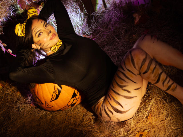 DorothyLane with tiger stripes on her legs and ass, black long-sleeve top, yellow headband, lying on hay, arms behind head, orange pumpkin with black eyes, dark room.
