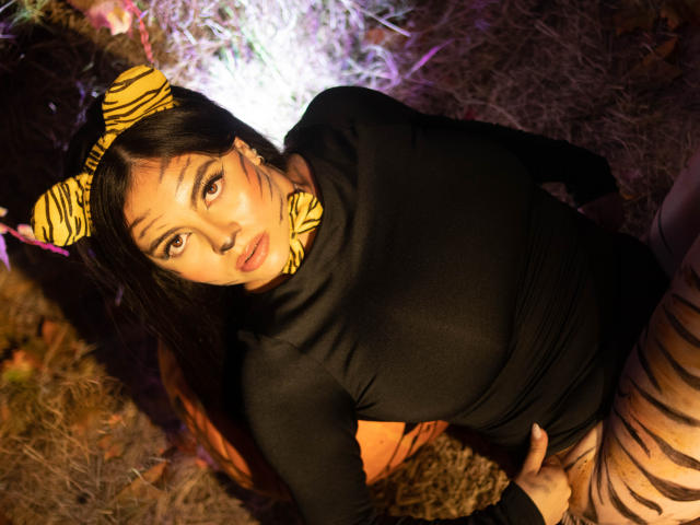 DorothyLane con una diadema a rayas de cebra y una blusa negra, mostrando sus pechos, con pantalones a rayas, mirando hacia arriba con una expresión de puchero, con una mano en la cadera, en un ambiente con poca luz..
