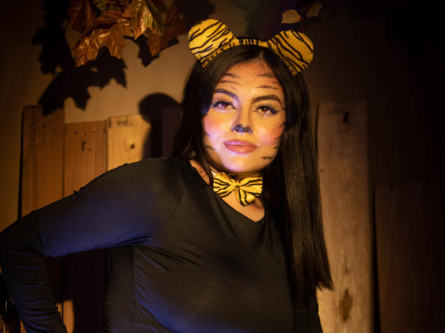 DorothyLane, con cabello negro, usando orejas de cebra, un aro en la nariz, y una blusa negra de manga larga, una corbata amarilla y marcas en la cara a rayas, de pie con las manos en las caderas, sonriendo..
