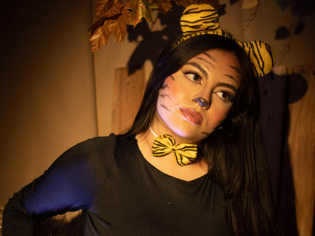 DorothyLane, com longos cabelos pretos, vestindo uma blusa preta, um laço borboleta listrado como um tigre e uma tiara combinando com orelhas. O rosto pintado com listras de tigre, posando com um sorriso divertido..