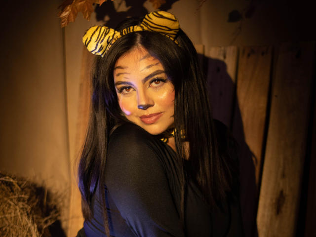 DorothyLane with long black hair, wearing a zebra headband and black top, has shiny makeup and a cute smile, looking over her shoulder, in warm, dim lighting.