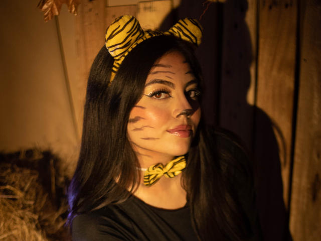 DorothyLane con lunghi capelli neri, una fascia e un collare a righe gialle a righe di zebra, finti segni a righe di zebra sulla guancia, che guarda la fotocamera, illuminazione marrone tenue, maglietta nera, sexy, carina..