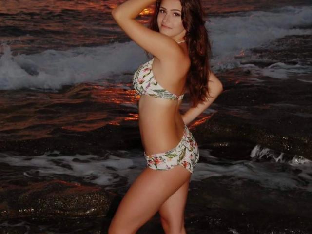 LiaMia-hot with curly brown hair, wearing a floral bikini, stands in dark, rocky water at sunset, hand on head, showing her ass and tits.