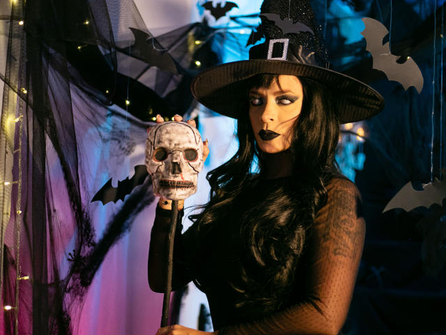 ChantalSex69 with black makeup, witch hat, and long black hair holding a skull on a stick, wearing a sheer black top, bat decorations in the background.