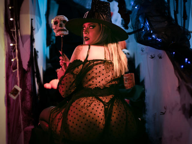 ChantalSex69 in a black polka-dot lingerie dress, holding a skull on a stick, wearing a witch hat, with blonde hair and a curvy butt, in a dark, spooky room.