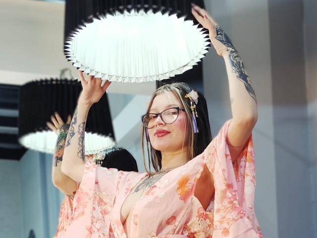 KoraSweet with glasses, pink floral robe, arms raised, holding a white, frilled lamp, tattoos on arms, blonde hair with purple streaks, red lipstick, looking up.