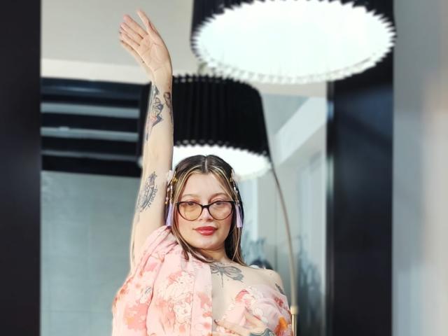 KoraSweet with glasses, pink floral top, and tattoos, raising arm, smiling, wearing pink headphones, has small tits, light skin, and brown hair, under a bright ceiling light.