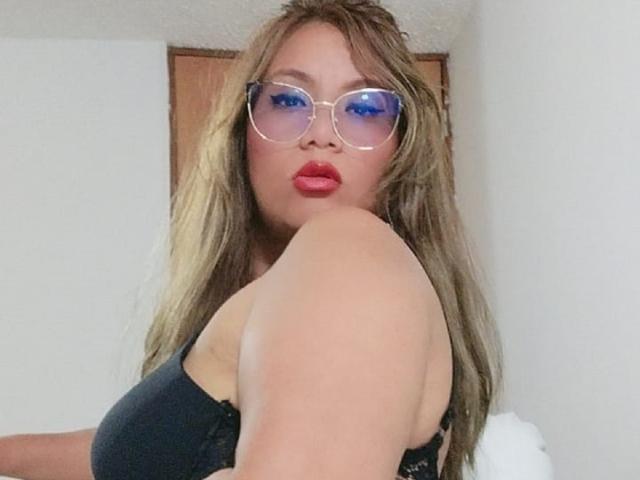 SamSexyest with blue eye makeup, red lipstick, and glasses, showing off her black bra-covered tits, with long brown hair and a slightly raised arm in a white room.