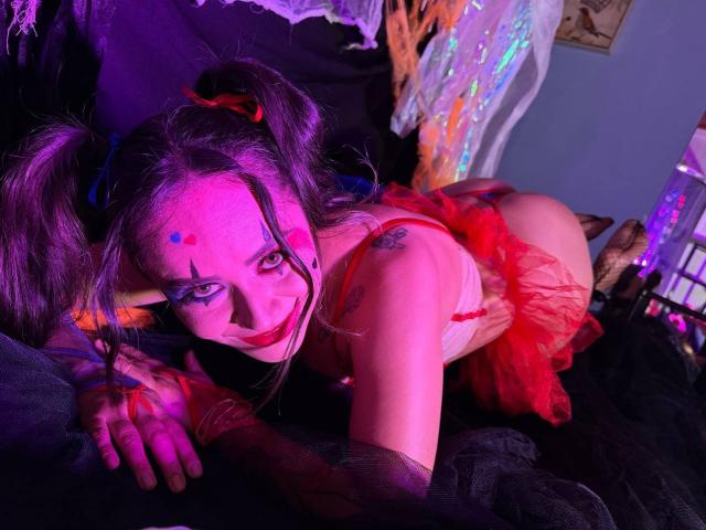 CamilaBardot with pink face paint, red lipstick, and blue eye makeup, in a red tutu, on all fours, showing her ass and tattoo, with purple and pink lighting.
