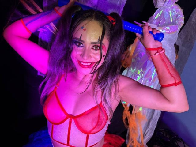 CamilaBardot with pigtails, red mesh bra, and red straps, holding a blue dildo, smiling, under purple and pink lights. Her tits are visible through the mesh.