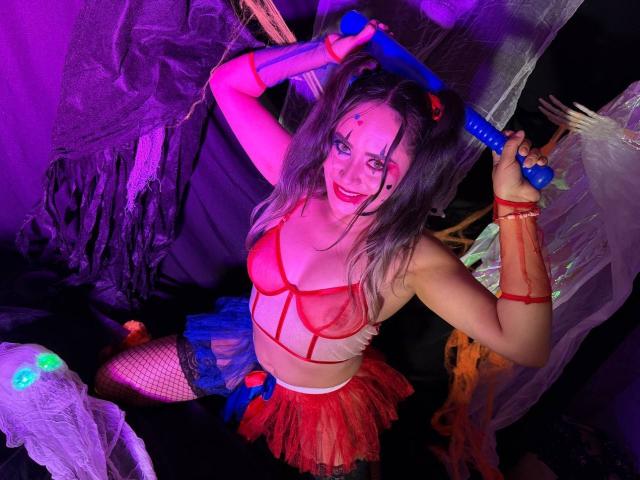 CamilaBardot with grey hair, red lingerie, fishnet stockings, and a blue dildo, holding it up with a playful smile, wearing a red tulle skirt, and purple makeup.