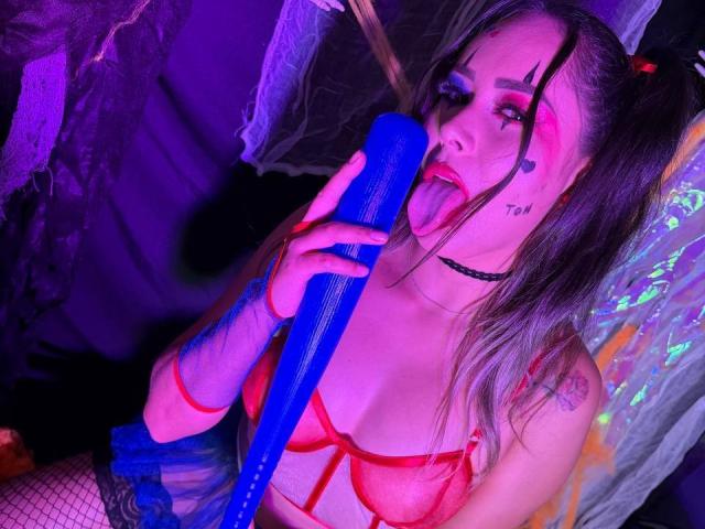 CamilaBardot with pigtails, tongue out, sucking a blue dildo, wearing red lingerie, fishnet arm, black choker, and tattoo on neck, under purple and blue lighting.