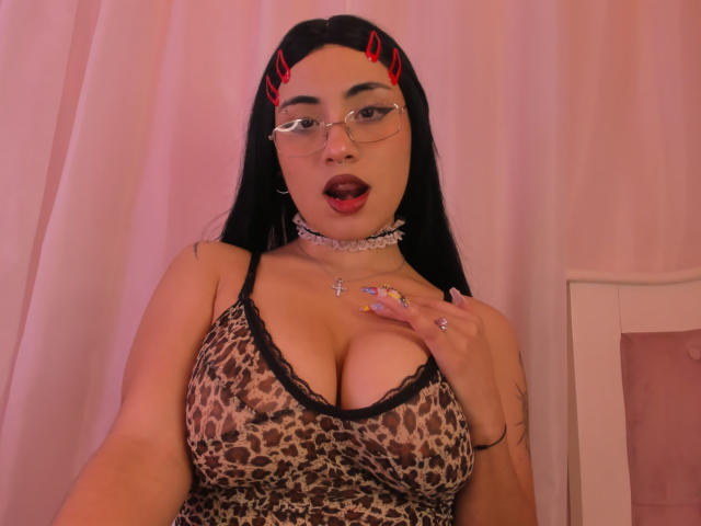 DiynaStar with black hair, glasses, and red hair clips, wearing a leopard print top with black lace, showing big tits, open mouth, pink choker, and tattoo on right arm.