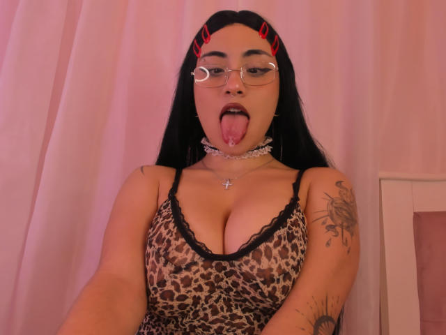 DiynaStar with long black hair, red devil horns, glasses, tongue out, leopard print dress, choker, cross necklace, tattooed arm, showing large tits, pinkish light, selfie.