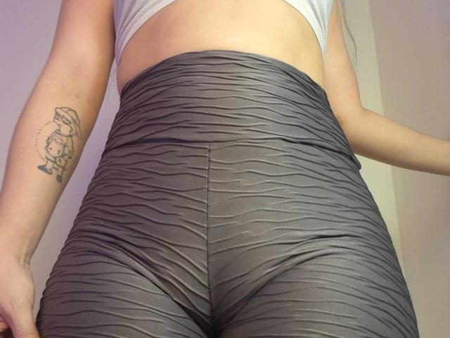 AmaiaSalvatoree's ass in tight, ruched gray pants, showing a tattoo of a cute cartoon cat on her right arm. She's wearing a white top, cut-off at the waist.