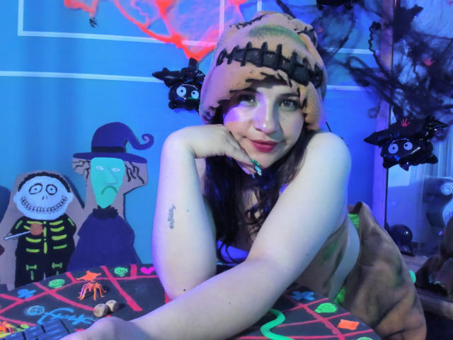 TaraTaylor, topless, with pale skin, wearing a pumpkin hat, resting her chin on her hand, smiling, with black cat toys and spooky decorations in a blue-lit room.