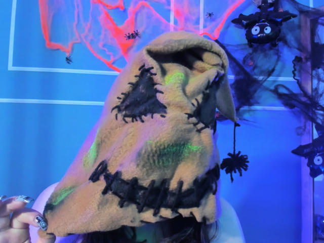 TaraTaylor wearing a creepy stitched-up, brown, fabric mask with green and black patches, black stitched eyes, and a black spider hanging from the nose. Blue and neon lights.