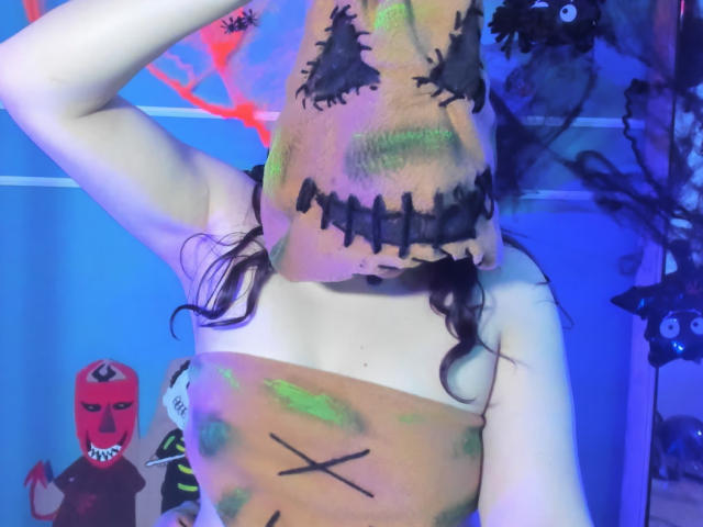 TaraTaylor with painted brown sack mask over head, showing stitched black eyes and black mouth. Paints on boobs and torso, long dark hair, arm raised. Blue lighting.